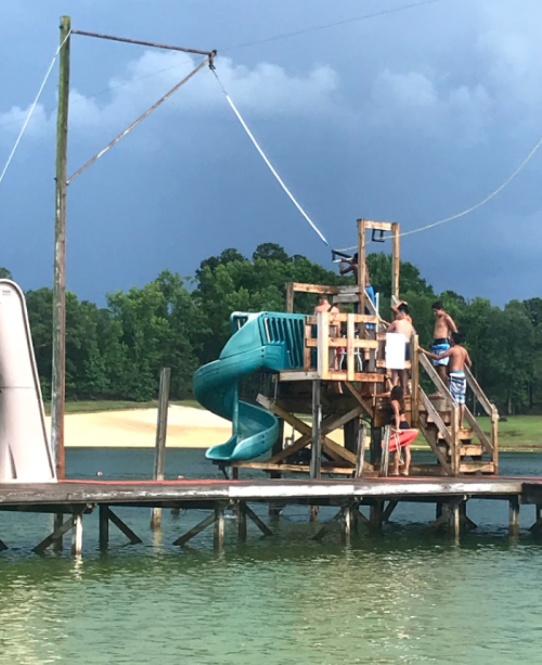 A group of people on a wooden dock with a water slide, preparing to jump into a lake under a cloudy sky.