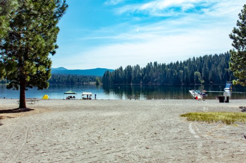 A serene lakeside scene with sandy beach, trees, and people enjoying the water under a clear blue sky.