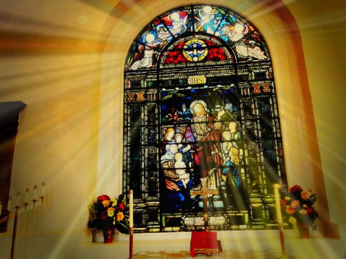 Stained glass window depicting religious figures, illuminated by sunlight, with flowers and candles in the foreground.