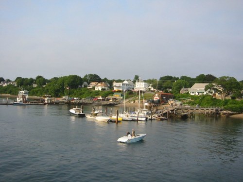 A serene waterfront scene with boats docked and a small motorboat on calm water, surrounded by lush greenery and homes.