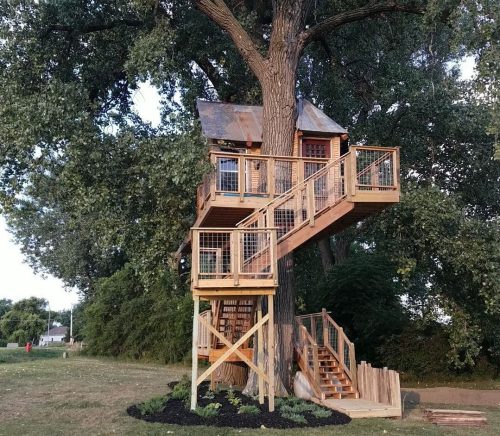 A wooden treehouse with a metal roof, elevated among trees, featuring a staircase and deck for access.