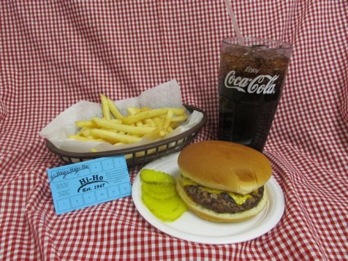 A plate with a hamburger, pickles, fries in a basket, and a glass of cola on a red checkered tablecloth.