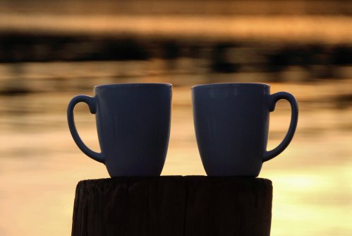 Two white coffee mugs sit on a wooden stump, with a blurred sunset reflecting on the water in the background.
