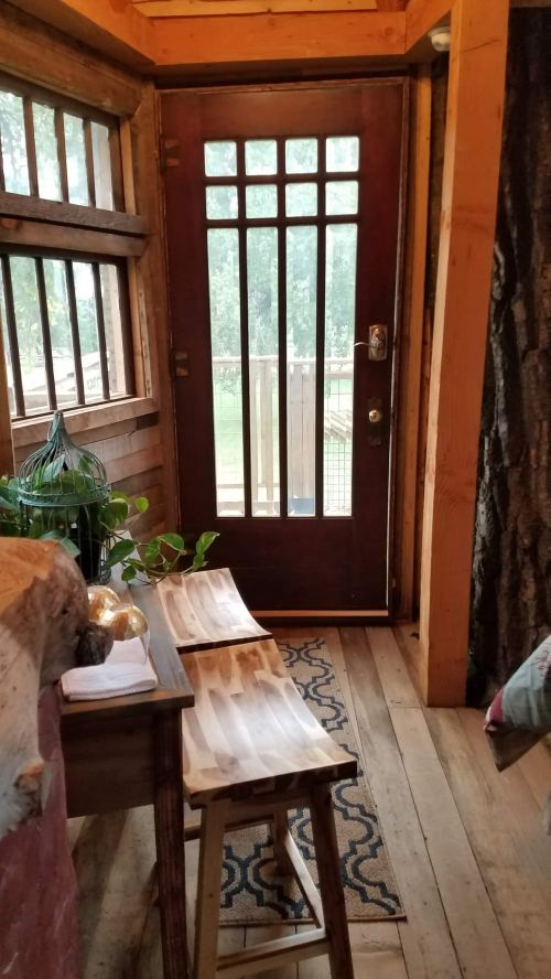 Cozy interior with wooden door, two stools, and a view of greenery through the window. Warm, rustic atmosphere.