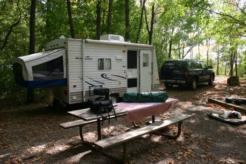 A camper trailer parked in a wooded campsite, with a picnic table and camping gear nearby.