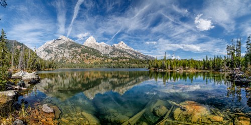 A serene lake reflects majestic mountains and a blue sky, surrounded by lush trees and rocky shores.
