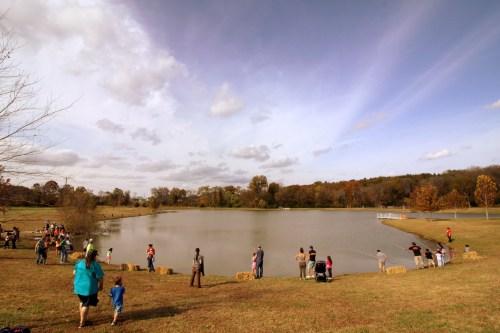 A group of people gather by a lake on a cloudy day, with trees and autumn colors in the background.