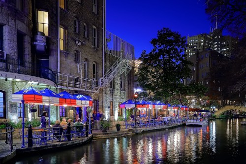 A vibrant riverside scene at night, featuring colorful umbrellas over outdoor dining along a canal, with city lights in the background.
