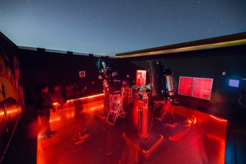 A dimly lit observatory with telescopes, red lighting, and people observing the starry night sky.