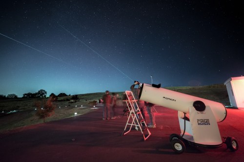 A group of people observing the night sky with a telescope under a starry sky, illuminated by red lights.
