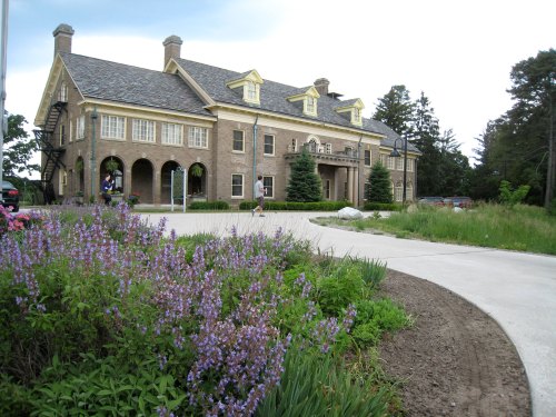 A large, historic brick house with yellow trim, surrounded by green grass and purple flowers, under a cloudy sky.
