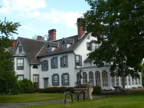 A large, historic white house with multiple gables, surrounded by greenery and featuring a porch and decorative fountain.
