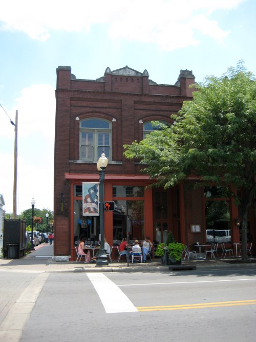 A red brick building with large windows, featuring outdoor seating and trees, on a sunny street corner.