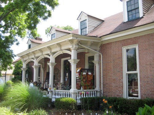 A charming brick house with a large porch, decorative columns, and lush greenery surrounding it.