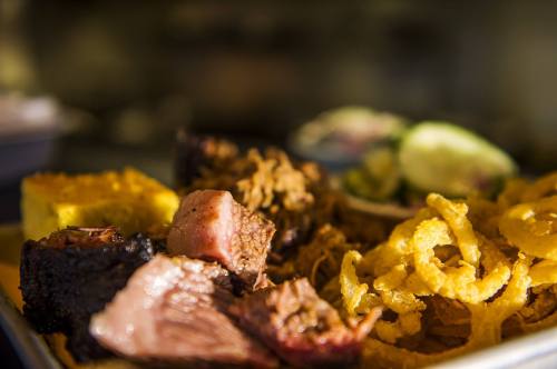 A close-up of a plate featuring various meats, cornbread, and crispy onion rings, with fresh vegetables in the background.
