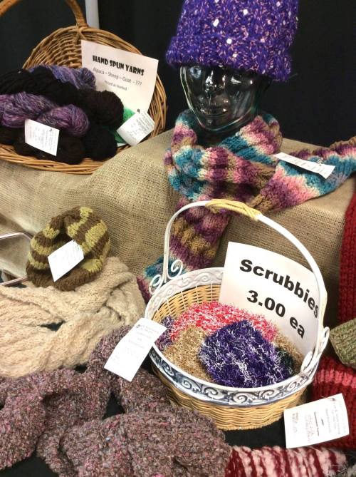 Display of handmade yarn products, including scarves, hats, and scrubbies, on a table with baskets and price tags.