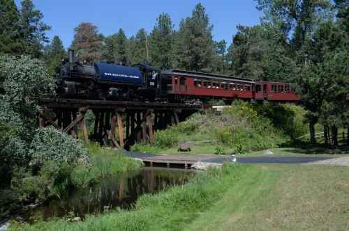 A vintage steam train crosses a wooden trestle bridge surrounded by trees and wildflowers.