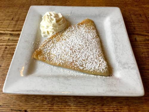 A triangular crepe dusted with powdered sugar, served with a dollop of whipped cream on a white plate.