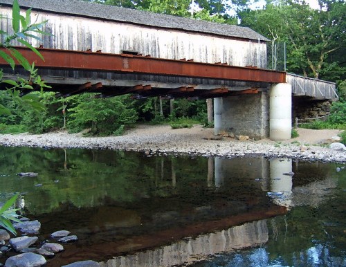 A rustic covered bridge spans a calm river, reflecting its structure in the water, surrounded by lush greenery.
