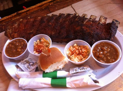 A plate featuring a large rack of ribs, sides of baked beans, coleslaw, and a bun, with utensils on the side.