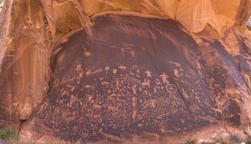 Ancient petroglyphs etched into a rock face, depicting various figures and symbols in a desert landscape.