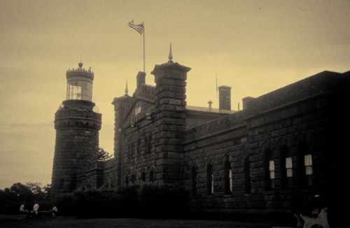Historic stone building with a tower and flag, surrounded by greenery, captured in a vintage style.