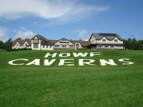 A grassy area with "HOWF CAVERNS" written in white, in front of a large, multi-story building under a blue sky.