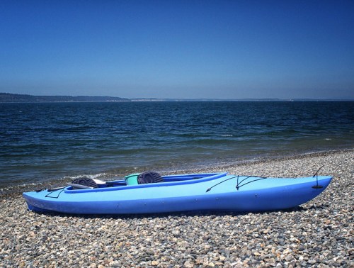 A blue kayak rests on a pebbly beach beside calm blue waters under a clear sky.