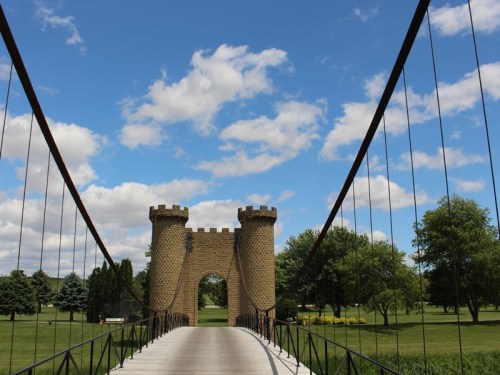 A suspension bridge leads to a castle-like structure under a blue sky with fluffy clouds and green trees.