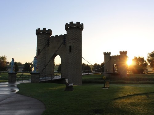 A castle-like structure with towers and a bridge, illuminated by a sunset, surrounded by green grass and trees.