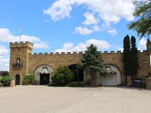 A stone castle-like building with a turret, arched entrances, and greenery under a blue sky with clouds.