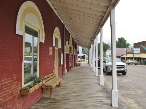 A wooden walkway alongside red brick buildings, with benches and parked cars under a cloudy sky.
