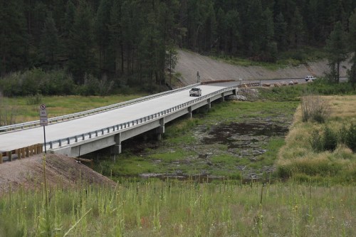 A bridge spans a small waterway, surrounded by greenery and trees, with a vehicle driving across it.