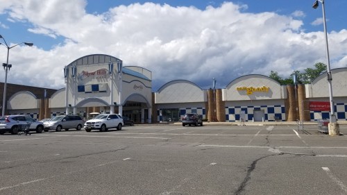 Exterior view of a shopping plaza with a large sign, featuring several stores and a mostly empty parking lot.