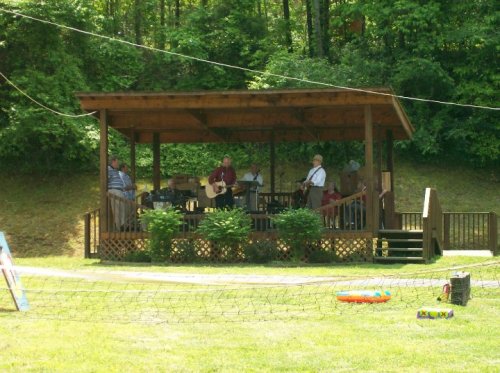 A wooden stage with musicians playing instruments, surrounded by greenery and a grassy area.