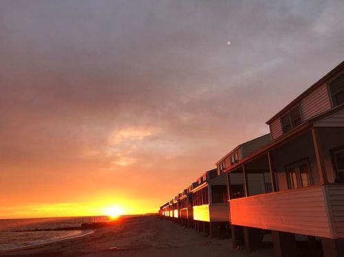 Sunset over a beach with silhouetted beach houses lining the shore, casting warm orange and yellow hues in the sky.