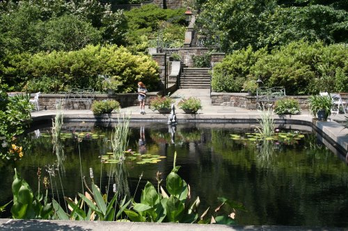 A serene garden with a pond, surrounded by lush greenery and flowers, featuring a child standing by the water's edge.