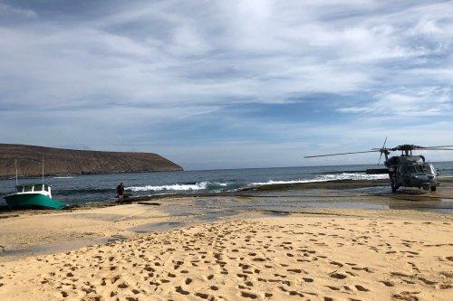 A beach scene with a helicopter and a small boat near the water, with footprints in the sand and a distant hillside.