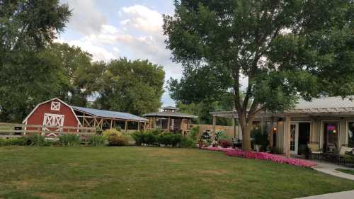 A scenic view of a red barn, a gazebo, and a house surrounded by green grass and colorful flowers under a cloudy sky.