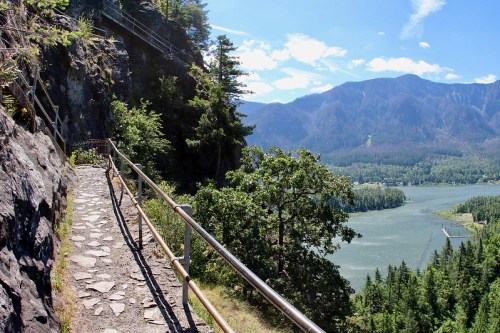 A scenic pathway along a cliff with a railing, overlooking a river and mountains under a clear blue sky.