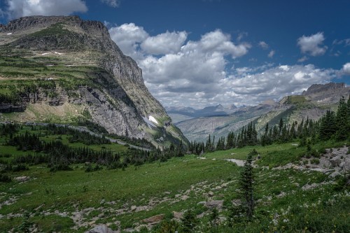 A scenic view of mountains and valleys under a partly cloudy sky, with lush greenery and rocky terrain in the foreground.