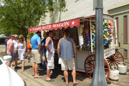 A busy hamburger wagon with people ordering food on a sunny day, surrounded by trees and colorful snack bags.