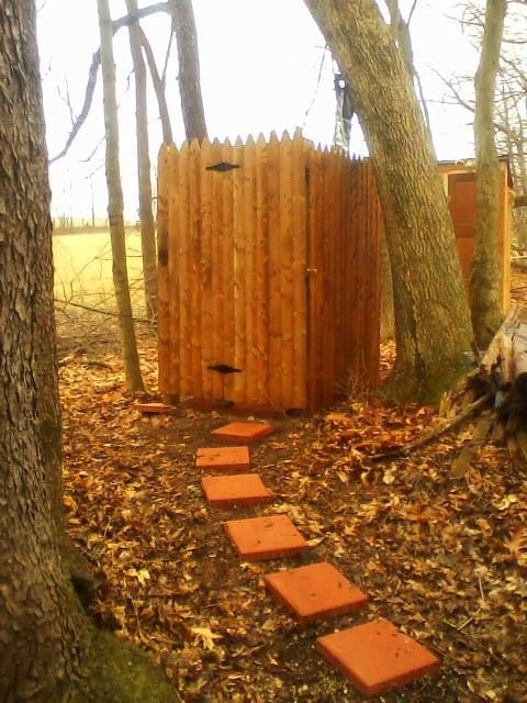 A wooden outdoor toilet with a pointed roof, surrounded by trees and a path of red stepping stones leading to it.