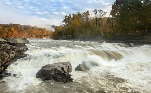 A rushing river flows over rocks, surrounded by trees with autumn foliage under a cloudy sky.