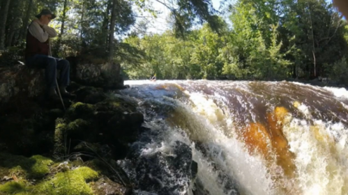 A person sits on a rocky bank near a rushing waterfall surrounded by lush green trees.