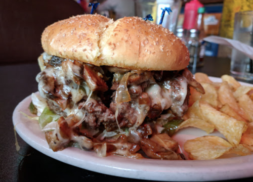 A large cheeseburger topped with melted cheese and mushrooms, served with a side of potato chips on a plate.
