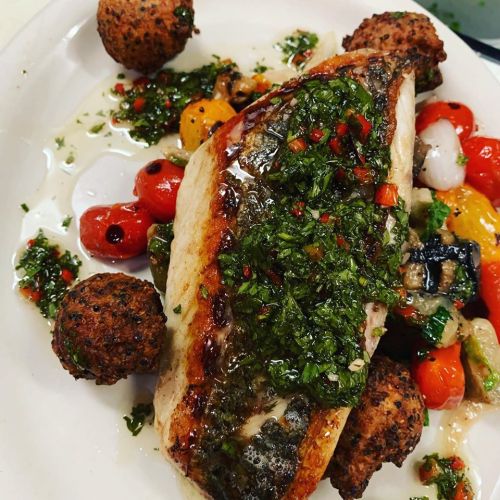 A plate of grilled fish topped with green sauce, surrounded by colorful roasted vegetables and falafel balls.