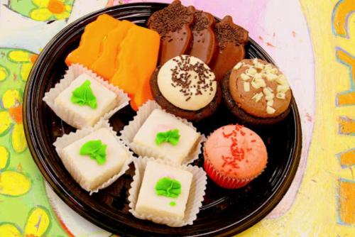 A black plate with colorful desserts: orange and chocolate treats, cupcakes, and small white confections with green accents.
