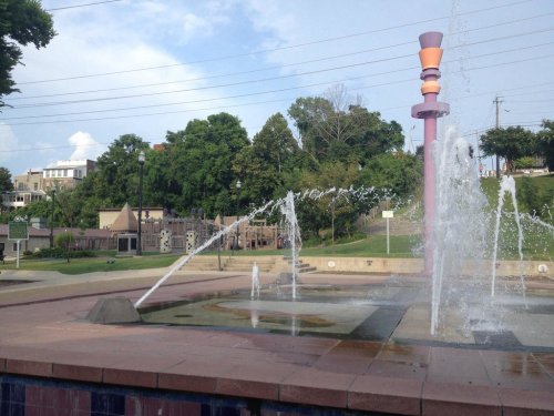 A colorful fountain sprays water in a park, with trees and playground equipment visible in the background.