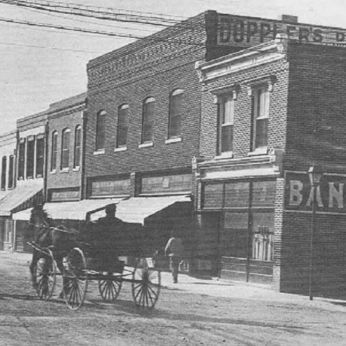 Black and white photo of a horse-drawn carriage in front of brick buildings with storefronts.